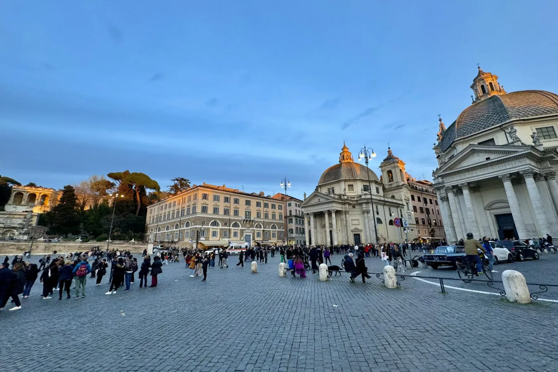 Piazza del Popolo in Rome with two domed churches and many people walking on the cobbled square at dusk.