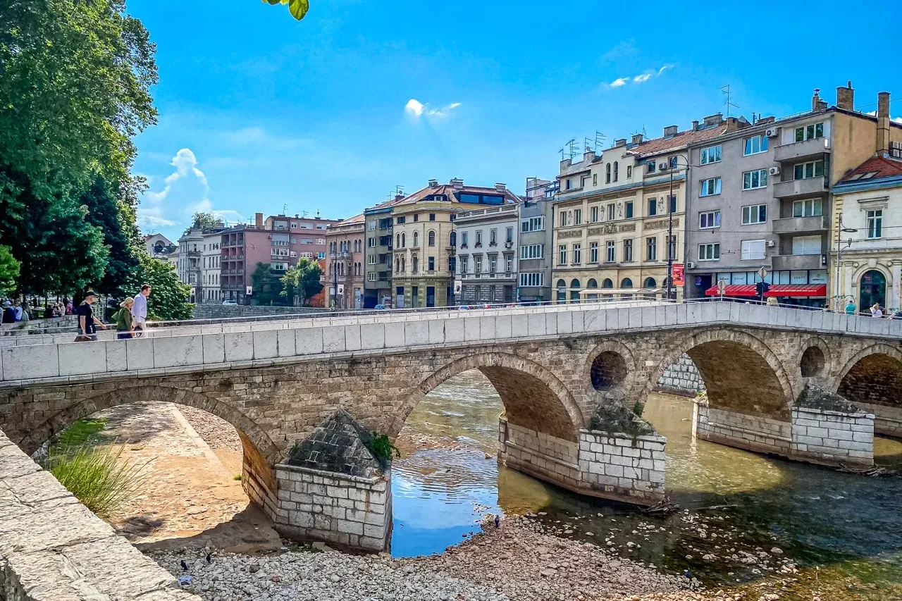 Stone arched bridge over a shallow river with historic buildings on the far bank under a blue sky, people walk on the bridge.