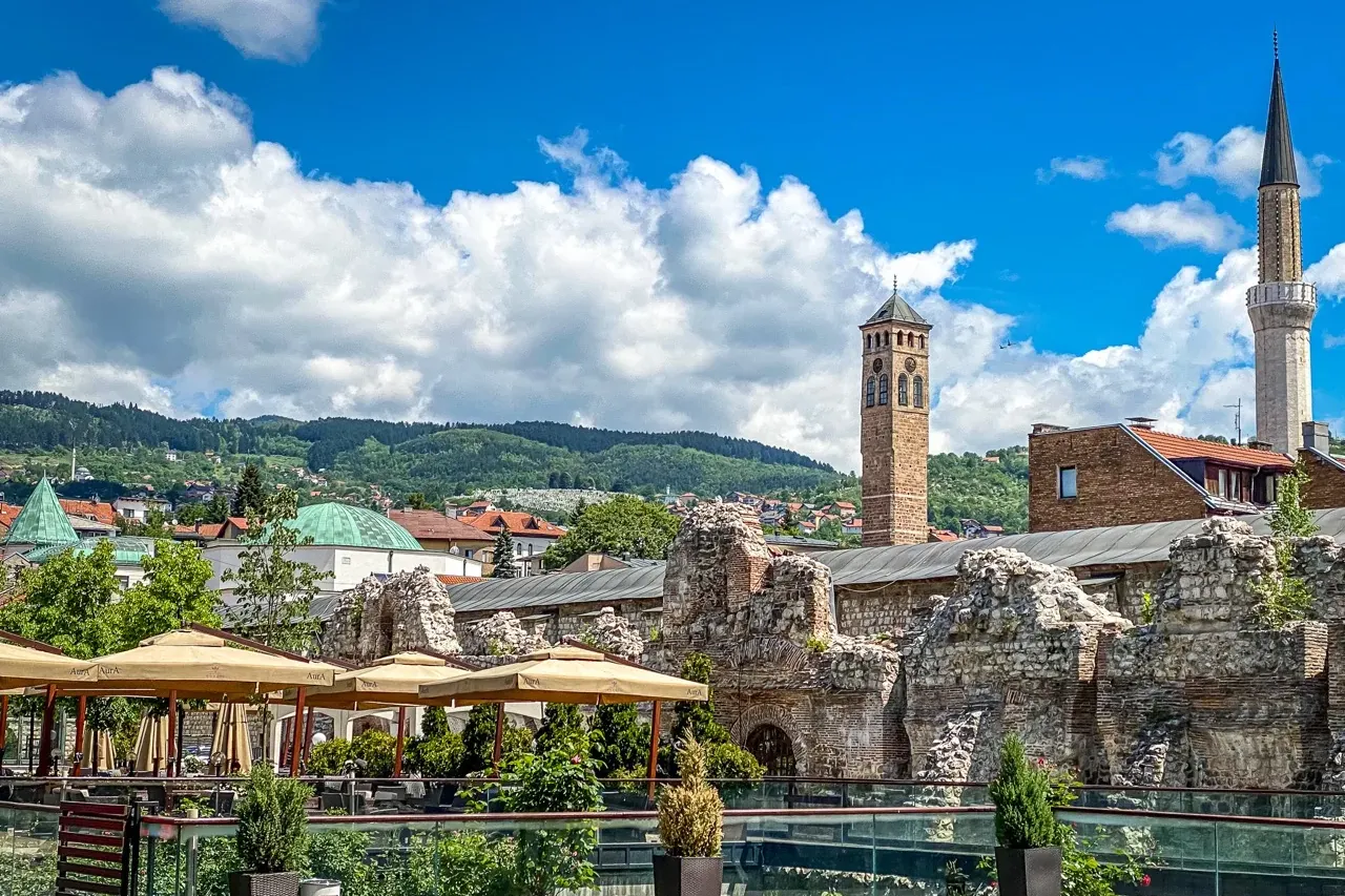 Historic Sarajevo cityscape with the Sahat Kula clock tower, a minaret, ancient ruins, and green hills under a blue sky.