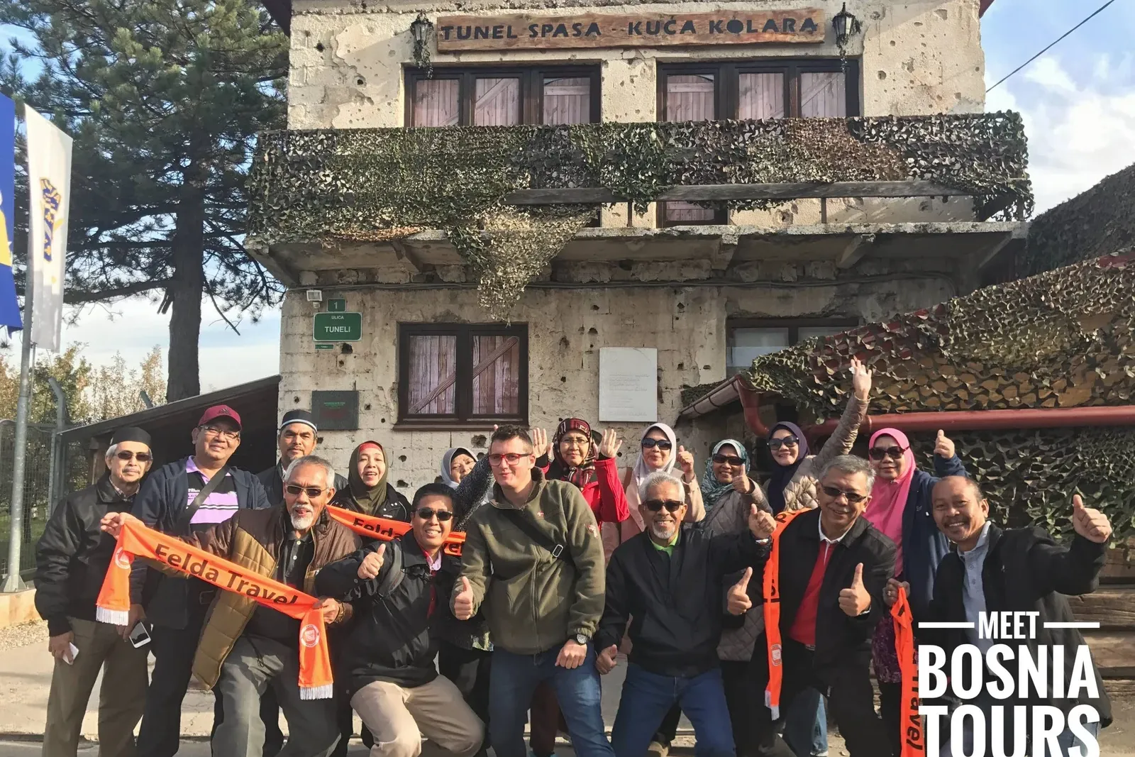 A group of diverse people stands smiling in front of the Tunnel of Hope entrance in Sarajevo, a building with visible bull...