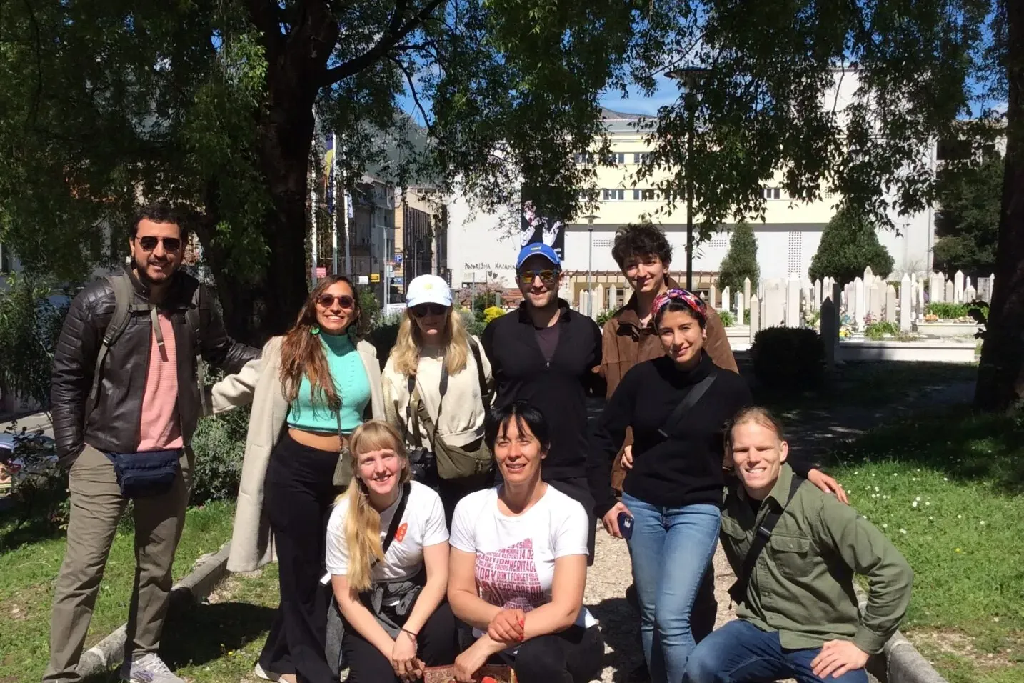 Group of 9 people smiling, posing for a photo outdoors under trees. In the background, a building and white gravestones ar...