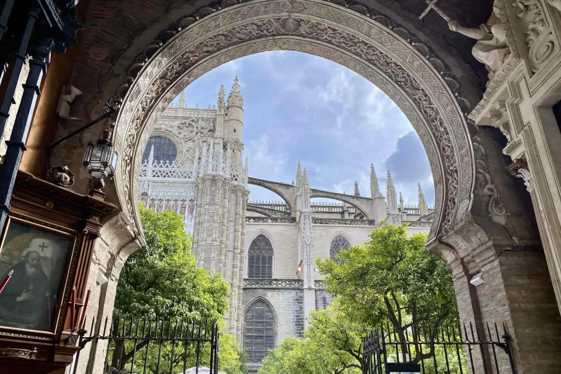 Gothic exterior of Seville Cathedral with spires and flying buttresses, framed by an ornate stone archway. Green trees vis...