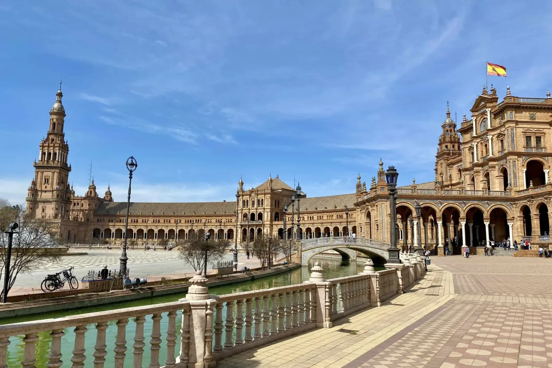 View of Seville's Plaza de España, showing the grand building with a tower, a canal with a bridge, and an ornate balustrade.