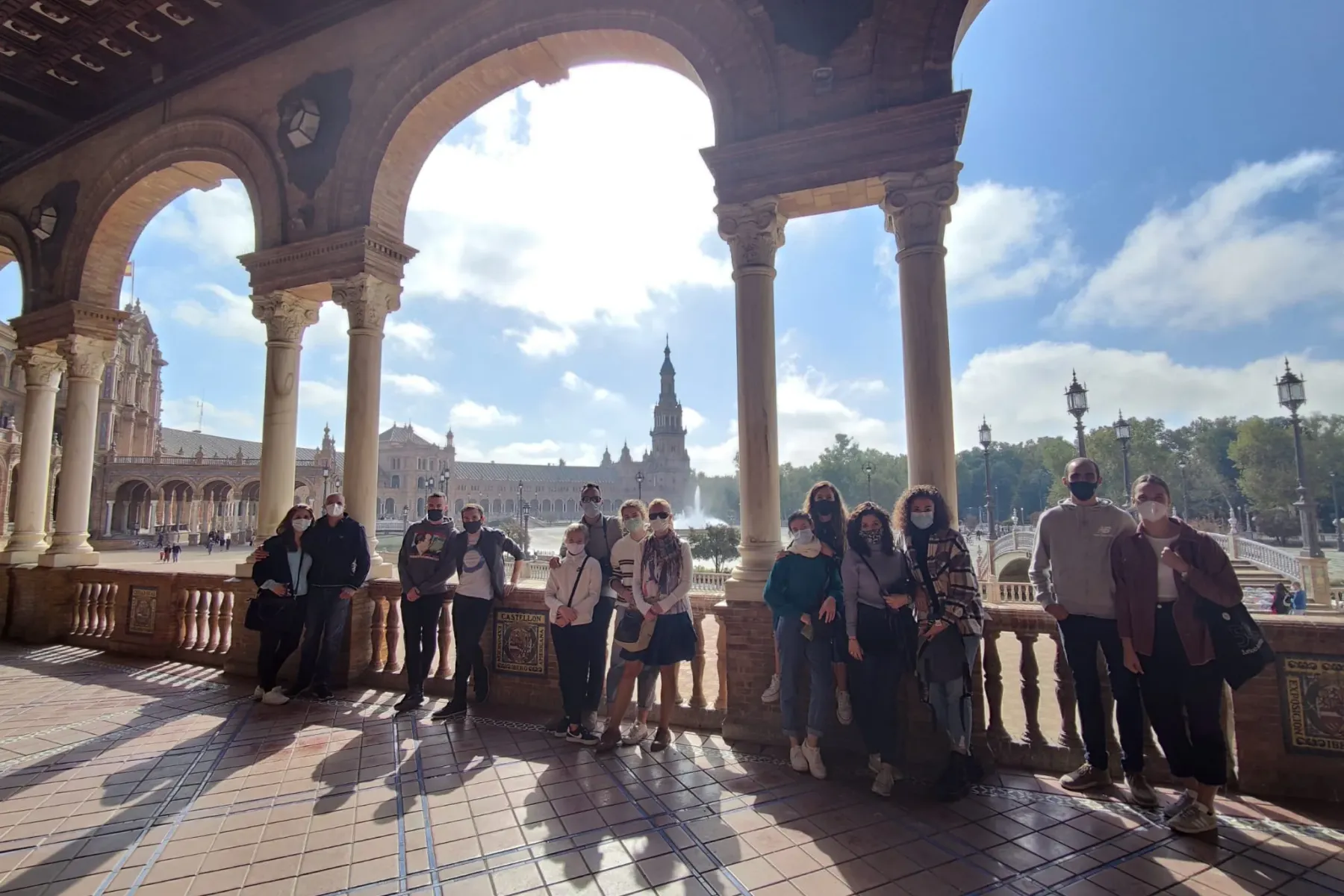 A group of people, many wearing masks, stand under a colonnade with arches, with Plaza de España and its tower in Seville ...