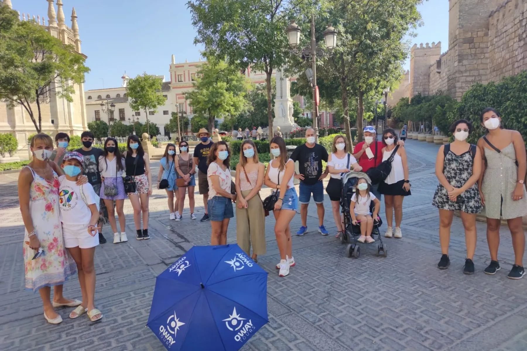 A group of people, many wearing masks, on a paved square in Seville. Historic stone walls are on the right, a building wit...