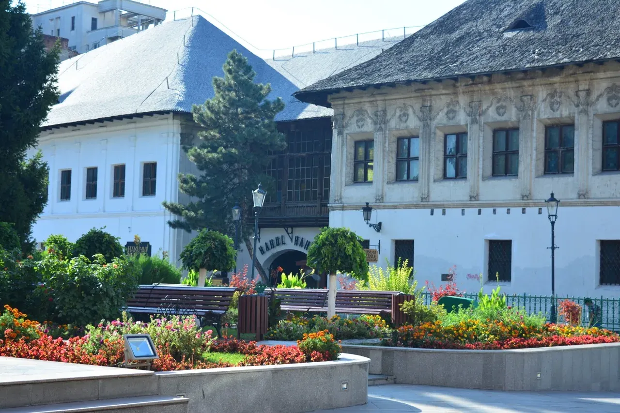 Historic buildings with textured roofs and a landscaped courtyard with colorful flower beds, benches, and trees under clea...