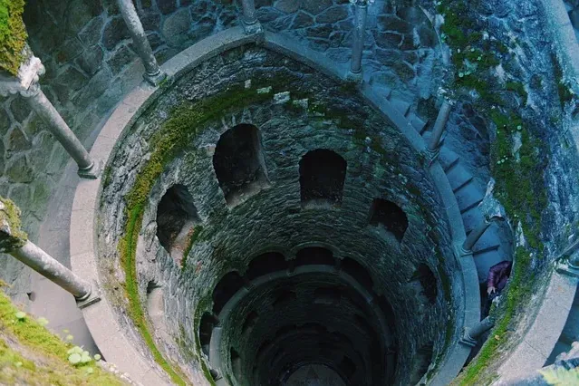 View looking down into a deep, moss-covered stone well with a winding spiral staircase. A person descends the steps.