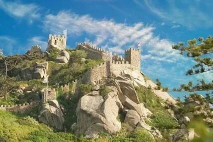 Ancient stone castle walls and towers crowning a steep, green, rocky hillside against a vibrant blue sky with scattered cl...