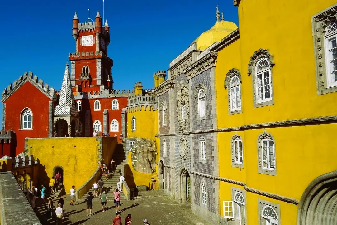 Exterior view of Pena Palace in Sintra with red and yellow sections, a clock tower, and tourists walking in the courtyard.