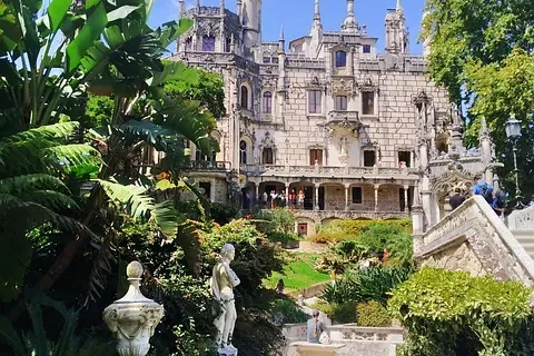 Ornate stone palace with turrets and balconies, surrounded by lush green gardens, palm trees, statues, and a path under a ...