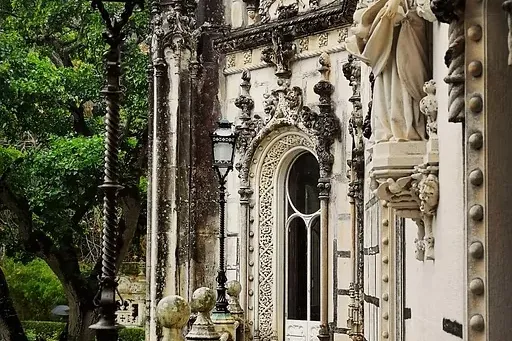 Ornate stone palace facade with sculptures, a decorative lamp post, and a wet, tiled walkway on a rainy day. Green trees v...