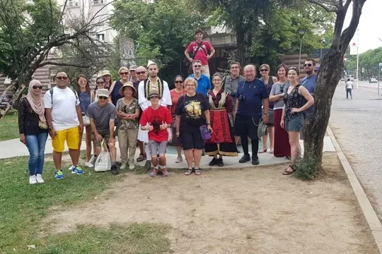 A diverse group of tourists, some in traditional Albanian attire, posing outdoors on a sunny day in Tirana. Trees and city...