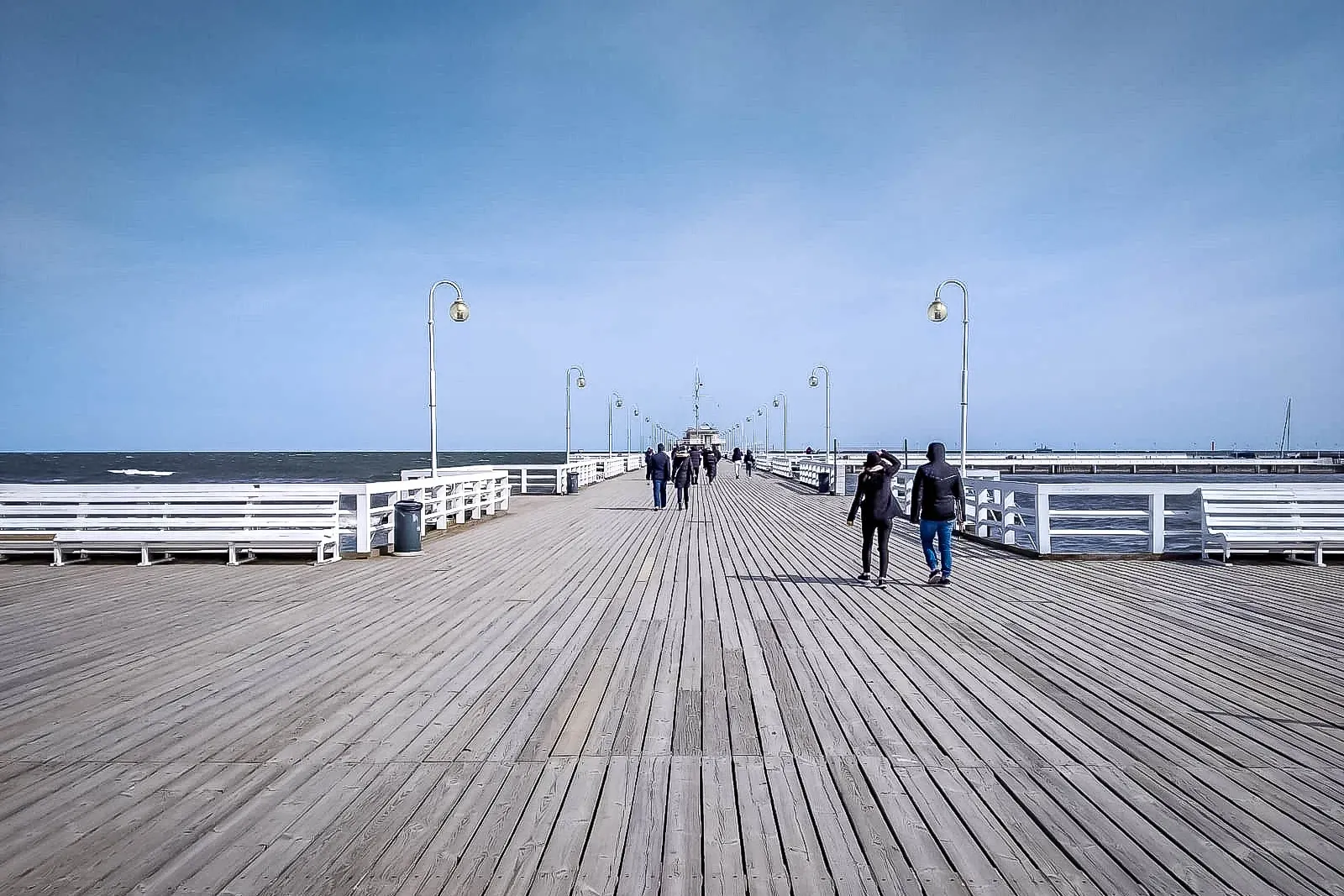 A long wooden pier stretching into the Baltic Sea, lined with white benches and streetlights, with people walking on it un...