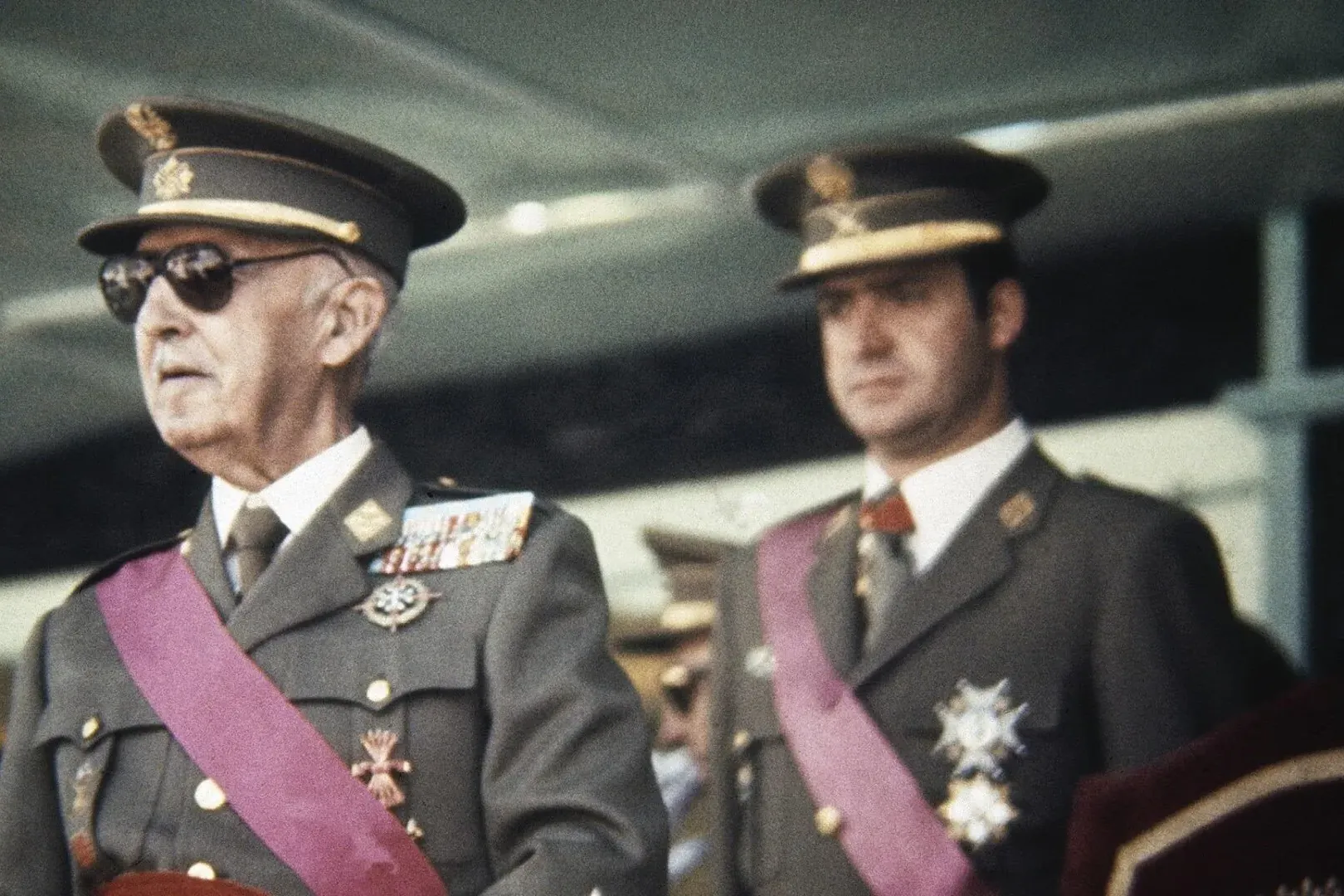 Francisco Franco, wearing sunglasses and military uniform with medals and a purple sash, with Juan Carlos I behind him.