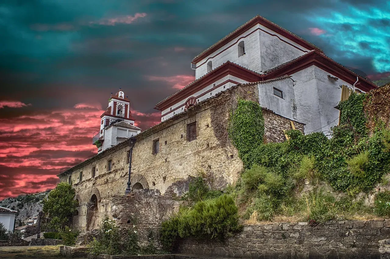Stone and white plaster building with red roof details and a prominent bell tower, against a dramatic sunset sky.