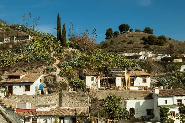 Whitewashed and rustic houses built on a steep, vegetated hillside under a clear blue sky. A narrow path winds through cacti.