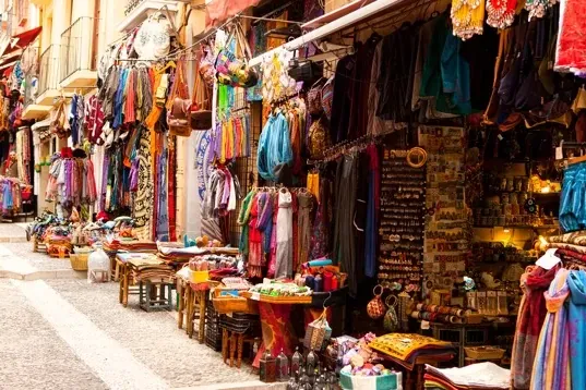 Narrow street in Granada lined with market stalls displaying colorful textiles, clothing, bags, and souvenirs. Buildings w...