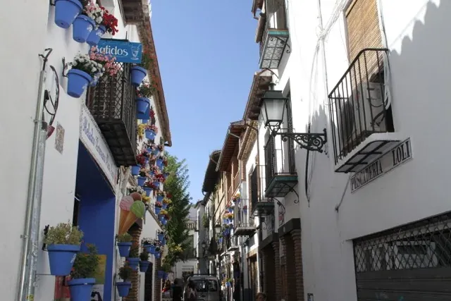 Narrow street in Granada with white buildings, blue flower pots, balconies, and a clear blue sky. A small car is visible.