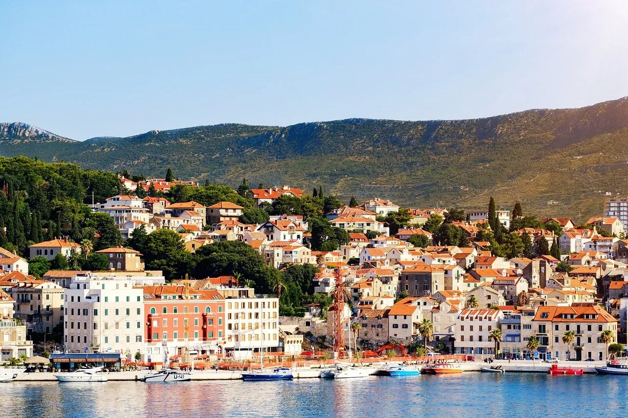 Coastal town with red-tiled buildings along a harbor, several boats docked, and lush green hills in the background under a...