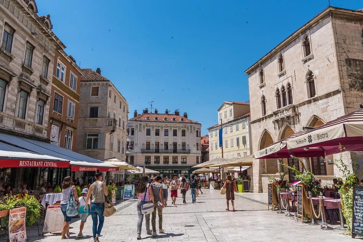 Crowded old town square in Split with stone buildings, outdoor cafes, and people walking under a clear blue sky.
