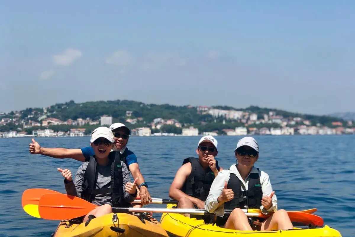 Four people in two yellow kayaks on blue water, smiling and giving thumbs up. A city with buildings is visible on a distan...