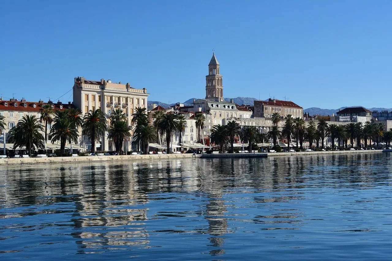 View of Split's Riva promenade with palm trees, historic buildings, the Cathedral bell tower, and calm sea reflecting the ...