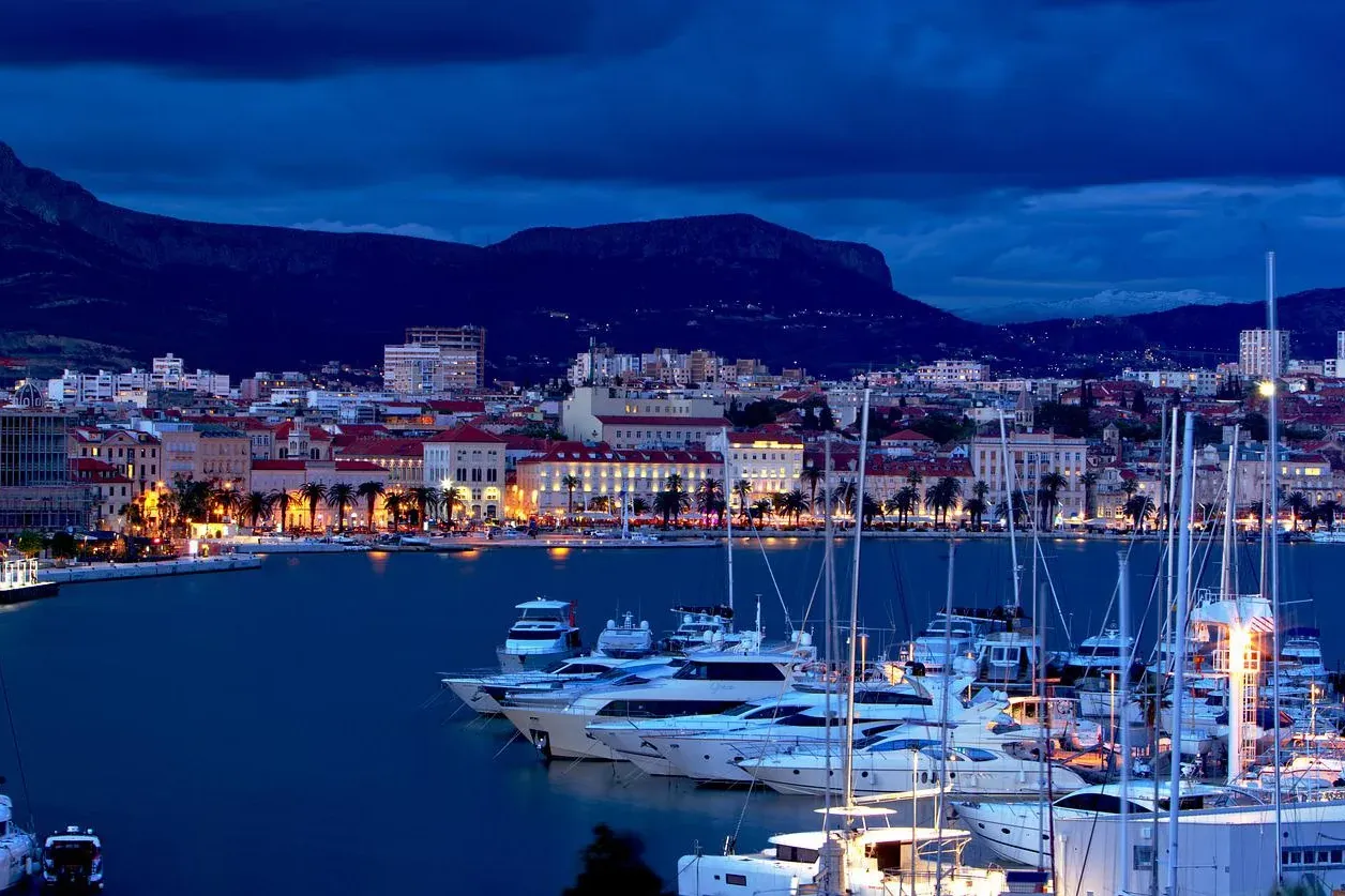Harbor at twilight with numerous yachts and boats, illuminated waterfront buildings, palm trees, and mountains under a dar...