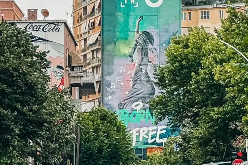 Street view with cars and green trees; a tall building with a large mural of a person and 'BORN FREE', topped by a domed r...