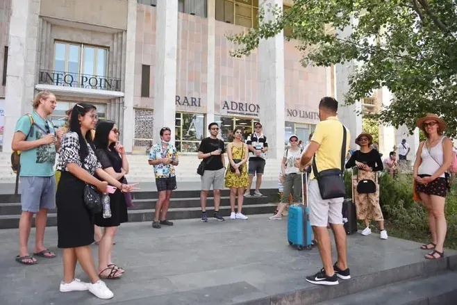 A diverse group of travelers listens to a male tour guide in a yellow shirt on a paved area in front of a modern building.