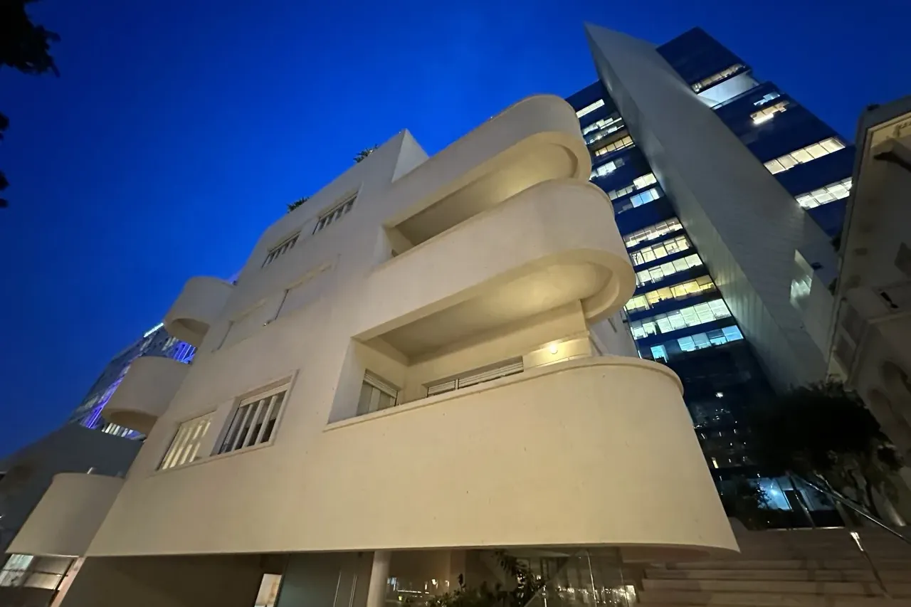 Cream-colored Bauhaus building with curved balconies under a dark blue sky, next to a tall modern glass skyscraper with il...