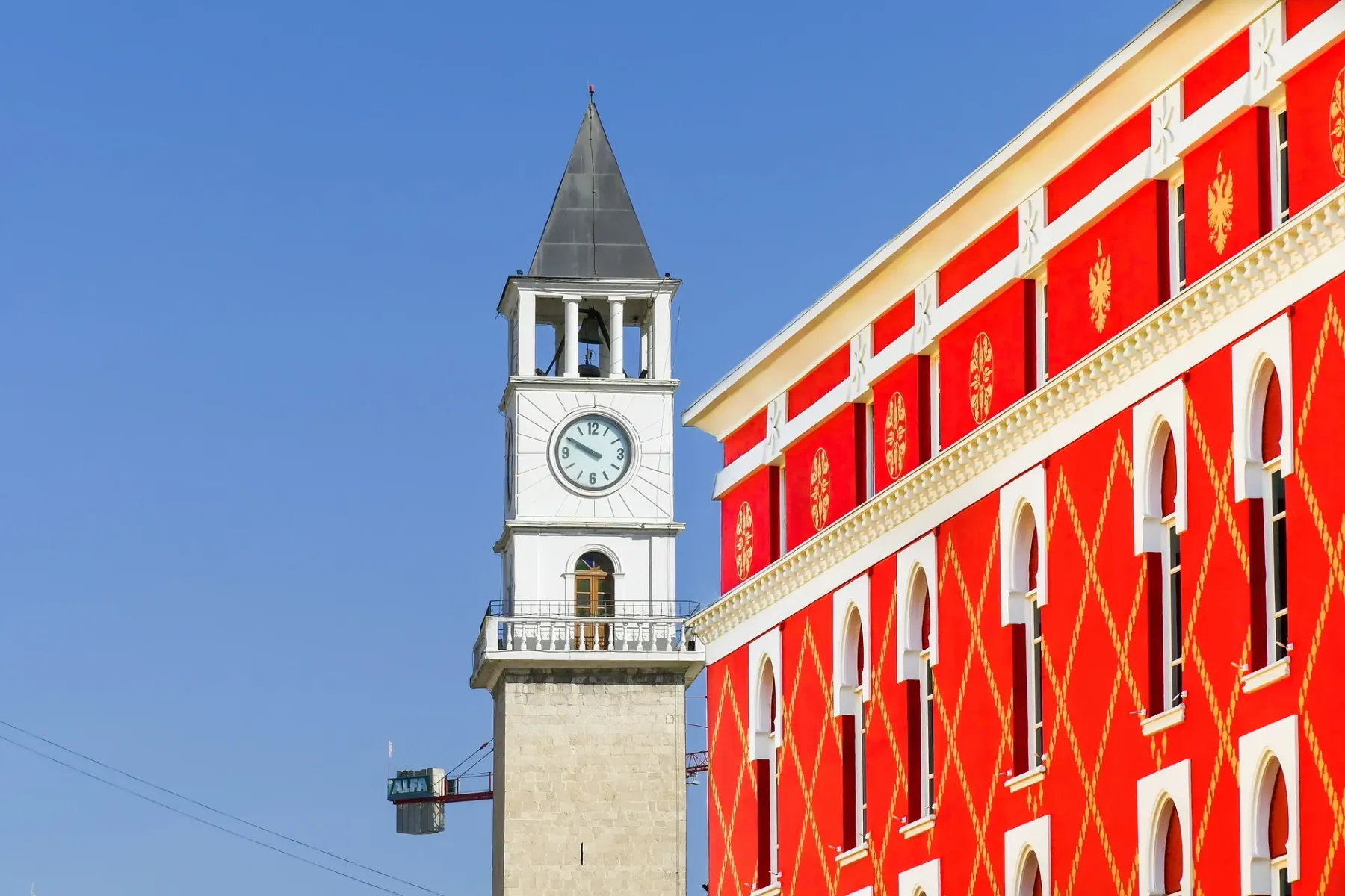 White clock tower with a black pyramidal roof and clock face, next to a red building with white arched windows and gold pa...