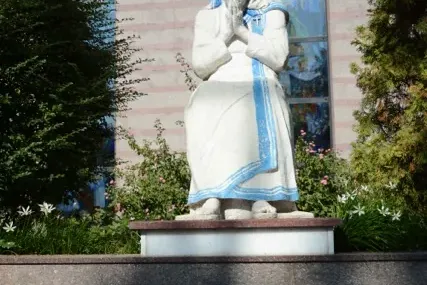 White statue of Mother Teresa with blue stripes, hands clasped, on a pedestal with a marble plaque in Albanian.