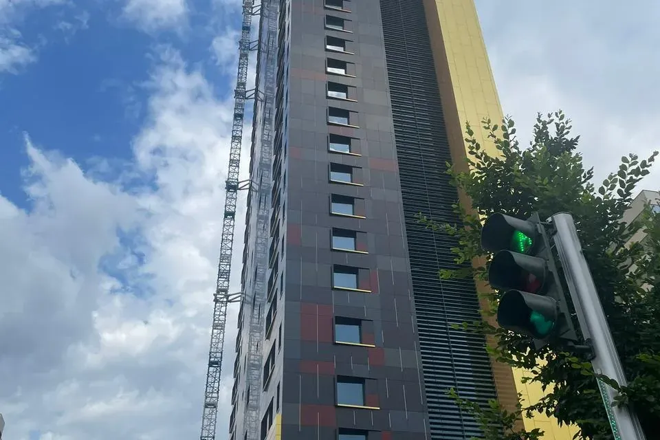 Tall modern building under construction with dark and gold facade panels, a large crane, and a green traffic light in Tirana.