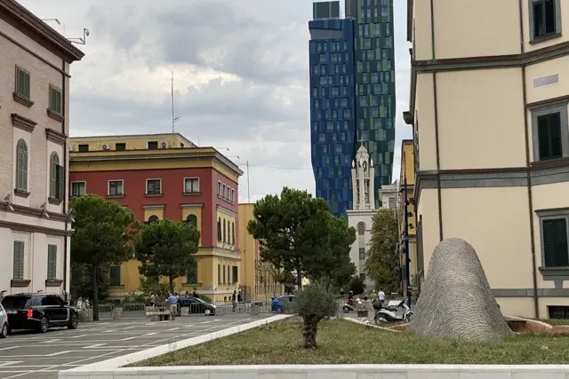Street view in Tirana with a modern blue and green skyscraper, traditional buildings, a church tower, and a unique grey sc...