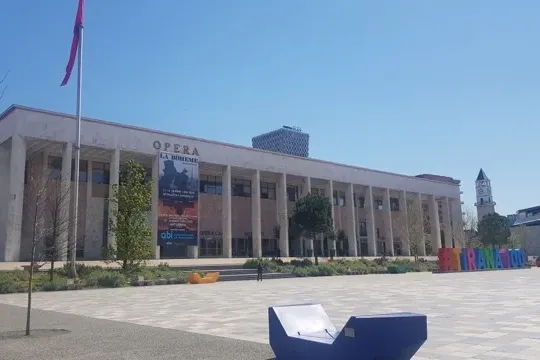 Large neoclassical building with 'OPERA LA BOHEME' banner, a square with modern benches, a clock tower and minaret in the ...