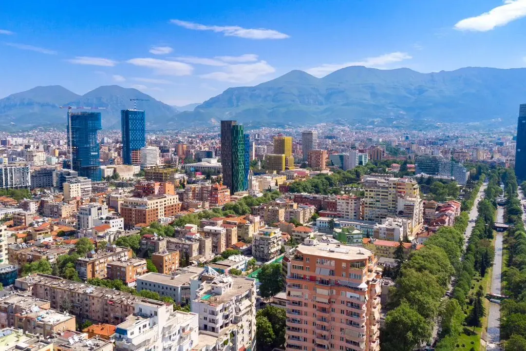 Aerial view of Tirana city with modern high-rise buildings, residential areas, a tree-lined boulevard, and mountains.