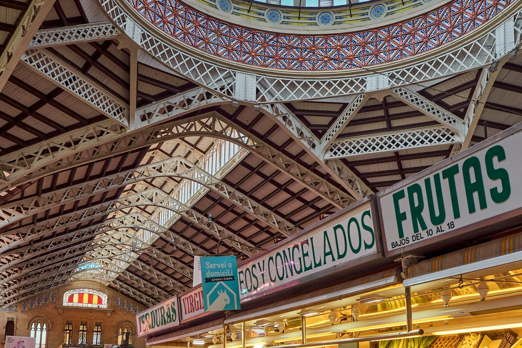 Market interior with fresh produce stalls, a metal roof structure, and an ornate, fruit-themed tiled dome ceiling with gla...