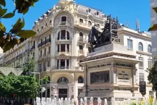 Ornate building and a monument with statues of Isabella and Columbus, next to a fountain, under a clear blue sky in Granada.