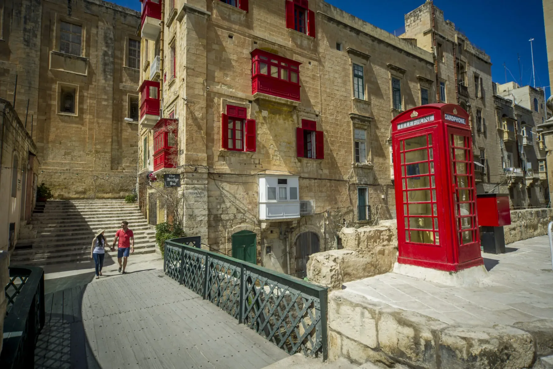 A couple walks over a bridge in Valletta, past buildings with red balconies and a red telephone booth.