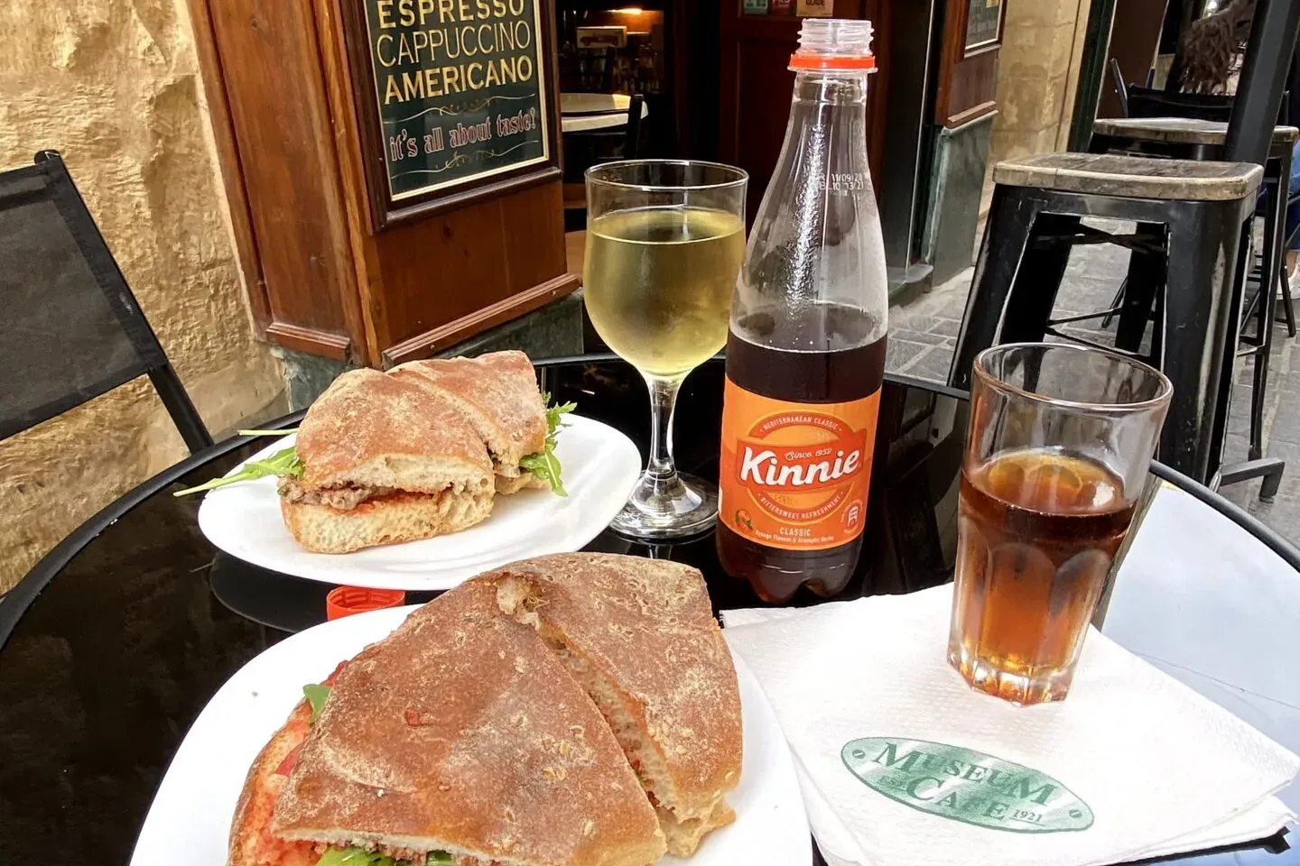 Outdoor cafe table with two sandwiches on ciabatta, a glass of white wine, a bottle of Kinnie, and a dark soft drink.