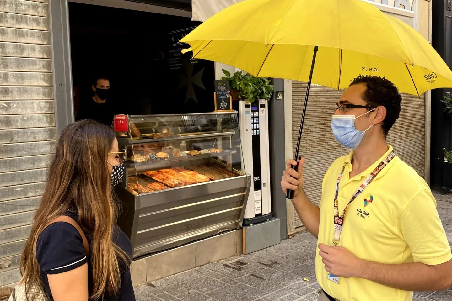 Man in yellow shirt holding umbrella speaks to woman in front of a 'Malta Pastizzi' shop with pastries. Both wear masks.
