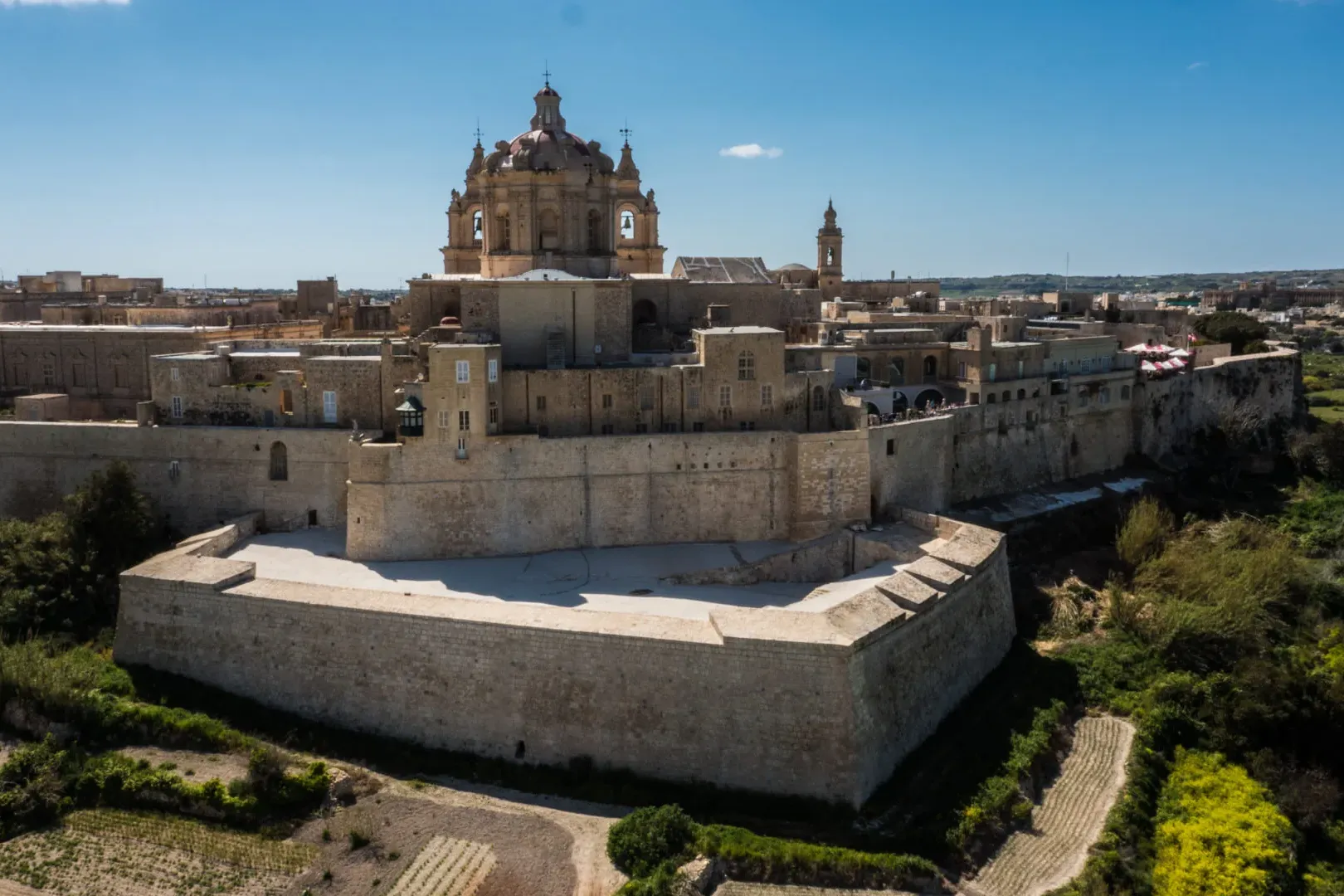 Aerial view of a historic fortified city with golden stone walls, buildings, and a large domed cathedral under a blue sky.