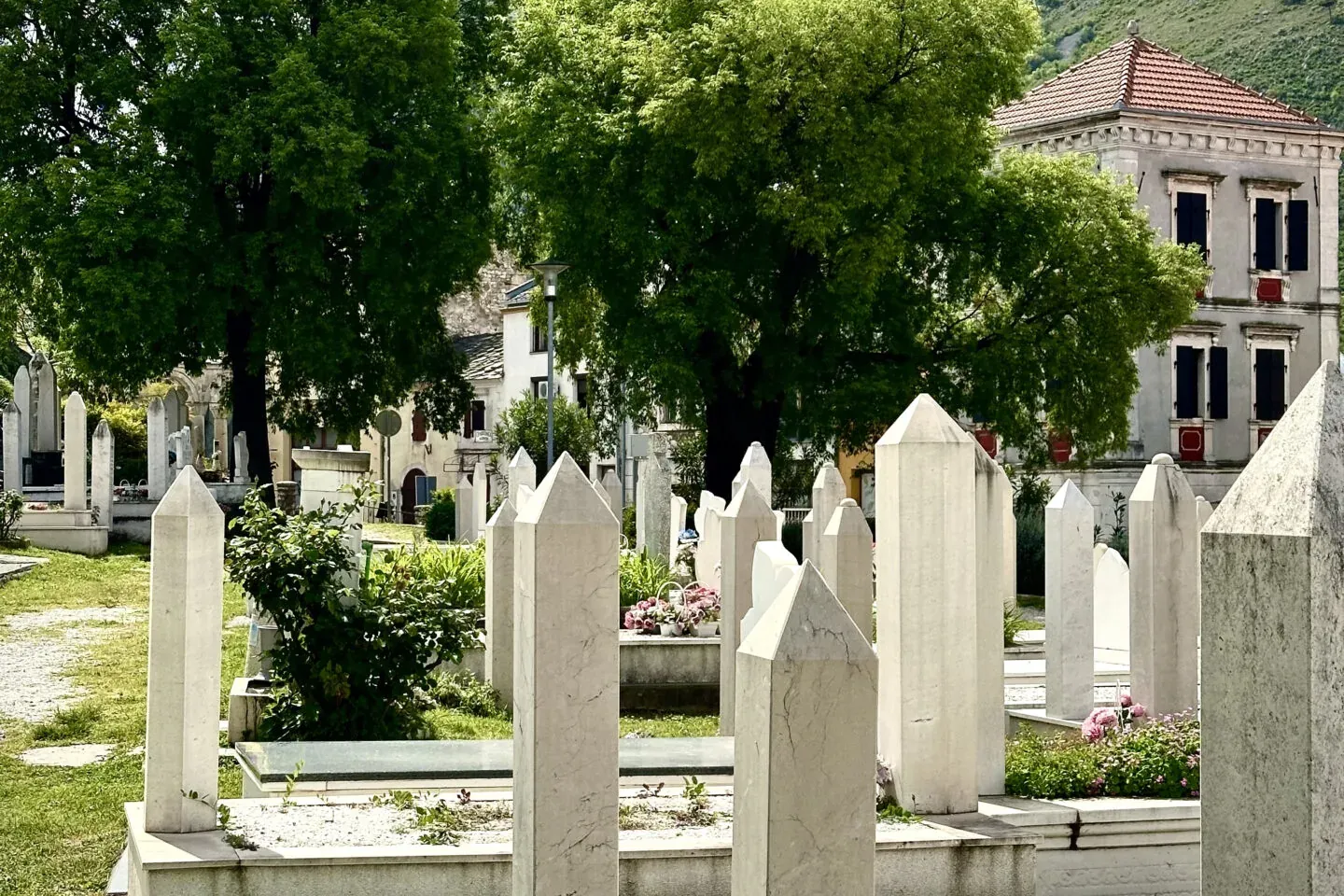 Angular white gravestones in a grassy cemetery, with green trees, an old building, and a distant hill topped by a white cr...