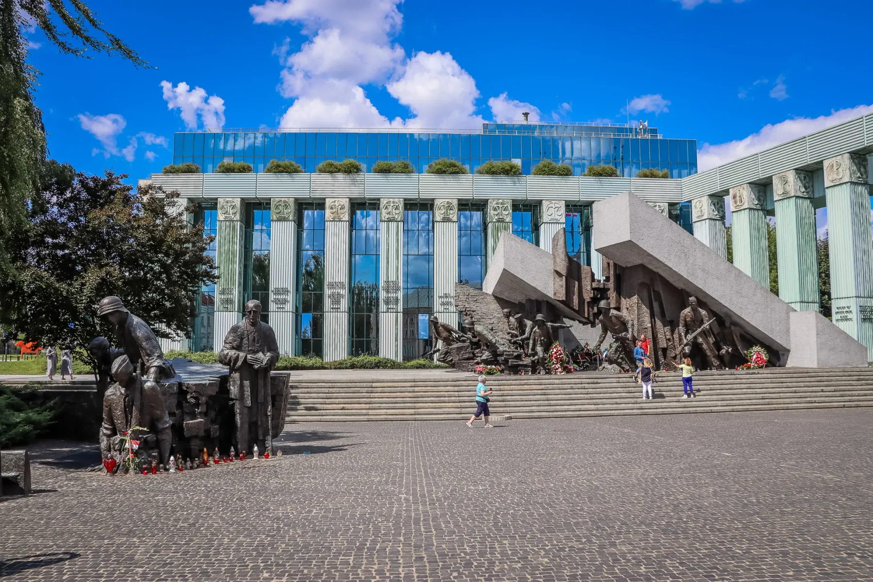 Bronze statues and a large concrete memorial stand on a cobblestone square in Warsaw. A modern building with a glass facad...