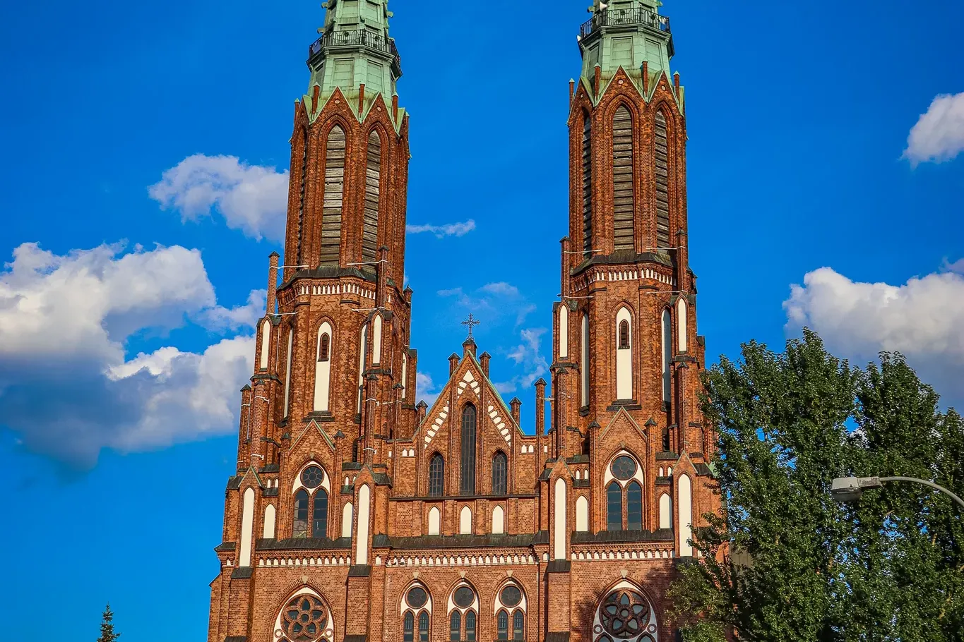 Tall red brick basilica with two green-capped spires reaching into a bright blue sky with white clouds. Green trees frame ...