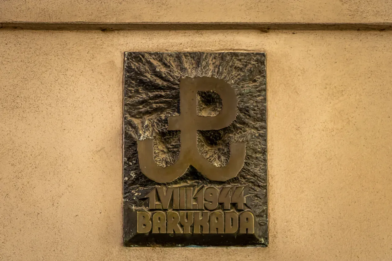 Bronze plaque on a light wall in Warsaw with the Polish Fighting A symbol, '1.VIII.1944', and 'BARYKADA'.