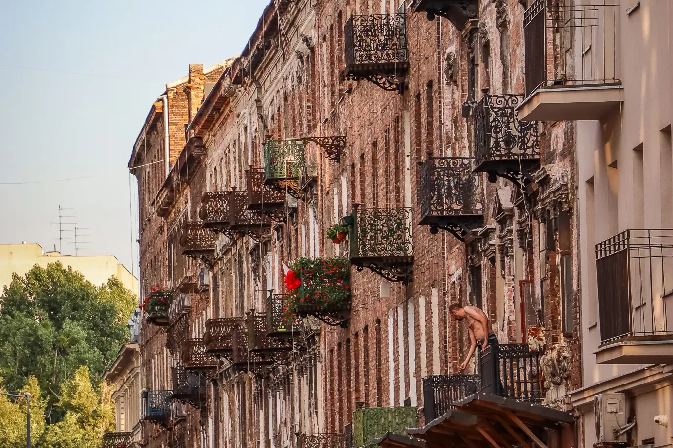 Street scene in Warsaw featuring old brick buildings with many ornate metal balconies. Parked cars and a red van are on th...