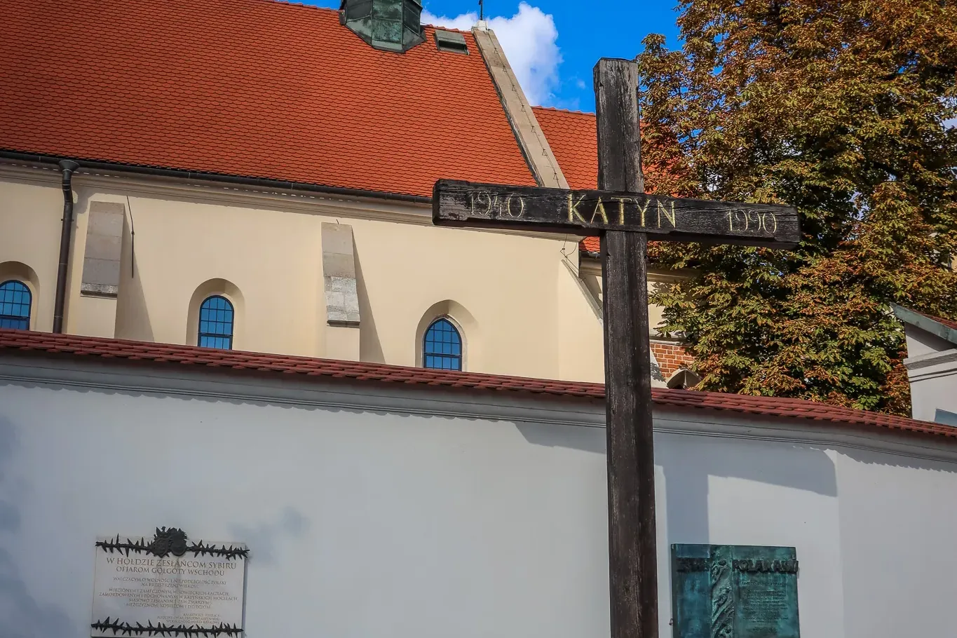 Large wooden Katyn Cross memorial with 'KATYN 1940 1990' inscription, plaques on a white wall, church roof, and spire.