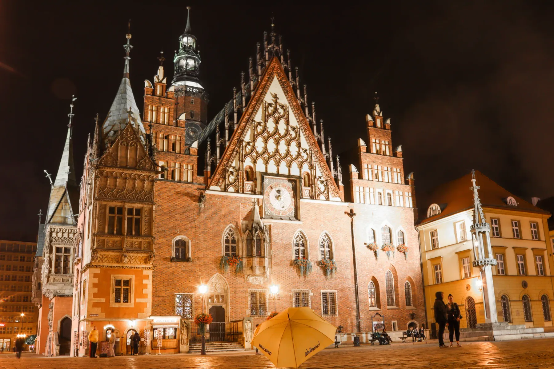 Illuminated Wroclaw Old Town Hall at night, showing its ornate facade, clock, and tall spire against a dark sky. A yellow ...