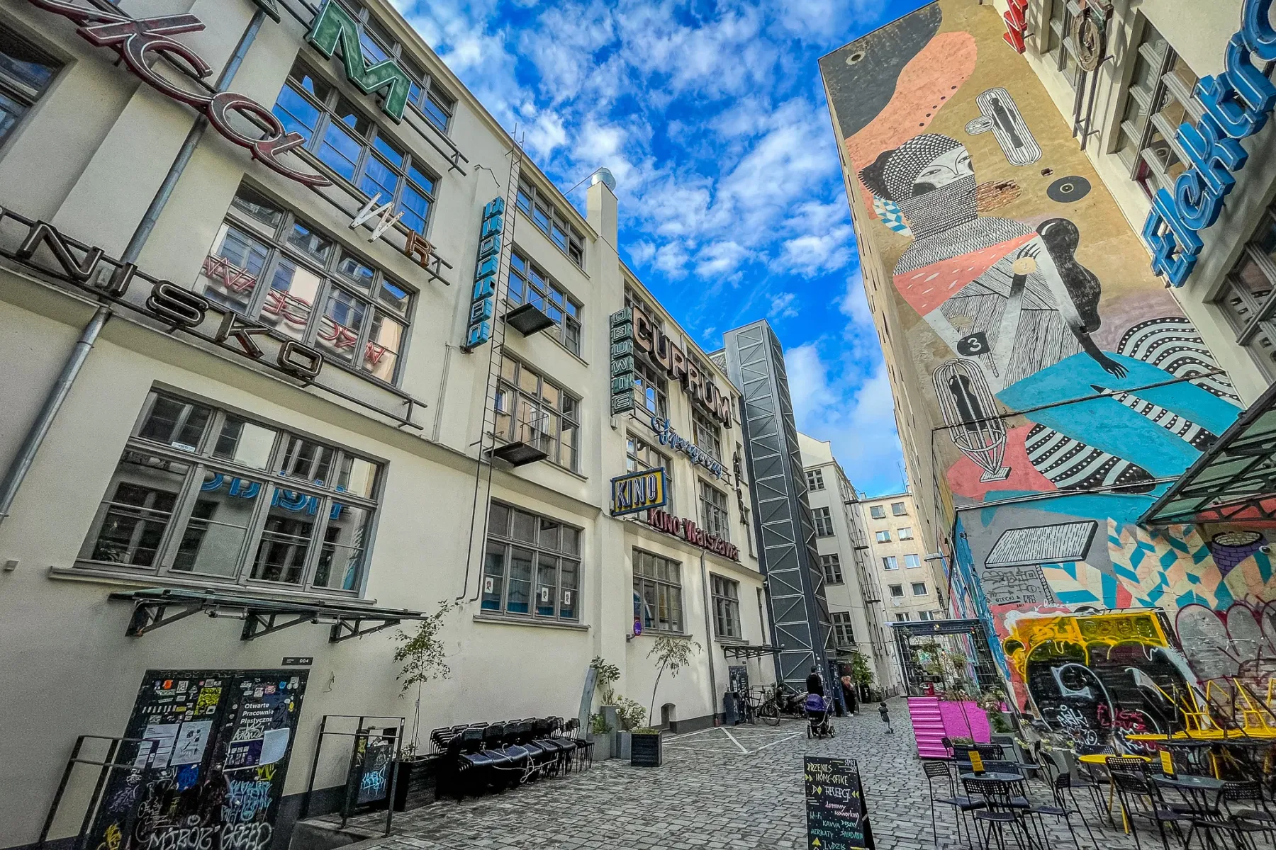 A narrow cobbled alleyway in Wrocław, flanked by a building with neon signs and another with a large abstract mural. Outdo...